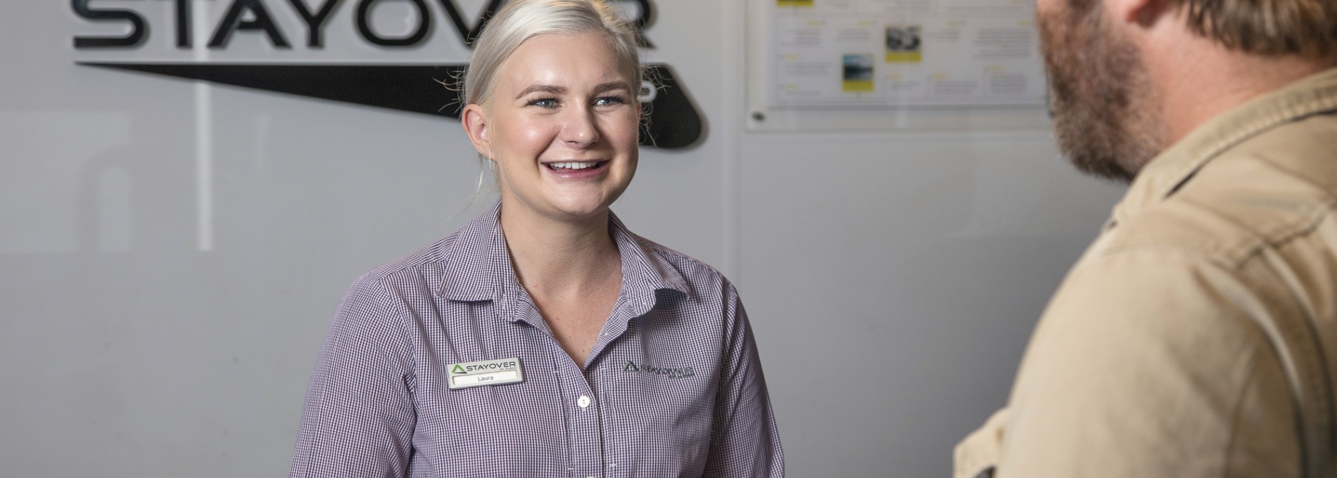 Woman Behind Front Desk Smiling at Man in Work Gear with Stayover Logo Behind Her